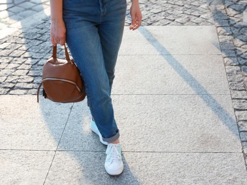 Pair of white sneakers on a lady wearing jeans and carrying a bag.