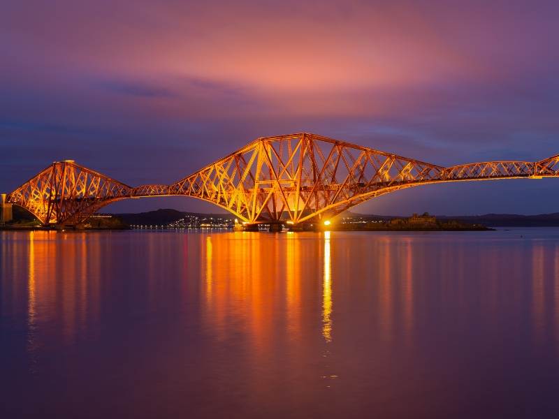 A large red bridge with reflections on the water and a purple night sky