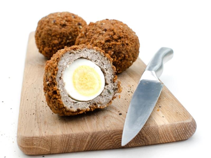 Scotch eggs on a wooden board with a knife. One of the eggs has been cut in half.