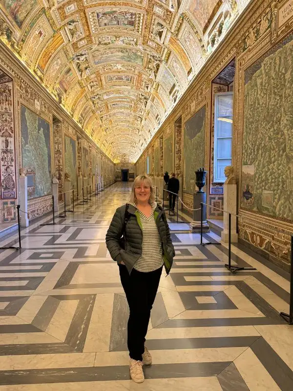 A woman standing at the end of a corridor with paintings on the walls and ceilings.